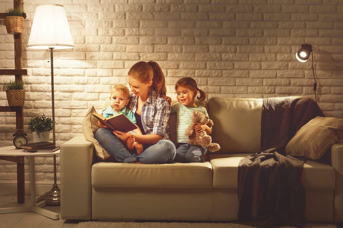 Mama, dieťa a dcéra si čítajú knihu na pohovke, vedľa nich svieti stojaca lampa